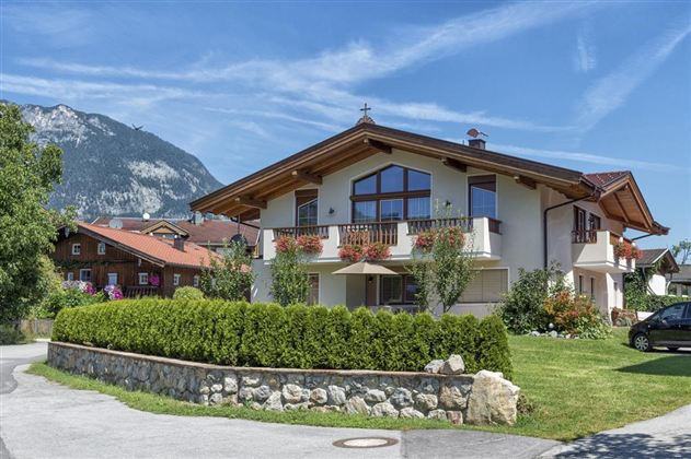 A beautiful, modern house with a well-maintained garden. In the background, mountains can be seen under a clear sky.