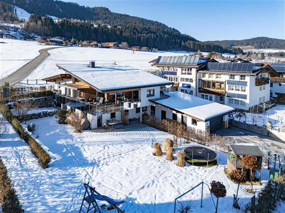 Ein großes Haus mit einem weiß verschneiten Garten und einem Trampolin. Im Hintergrund befinden sich weitere Gebäude und schneebedeckte Berge.
