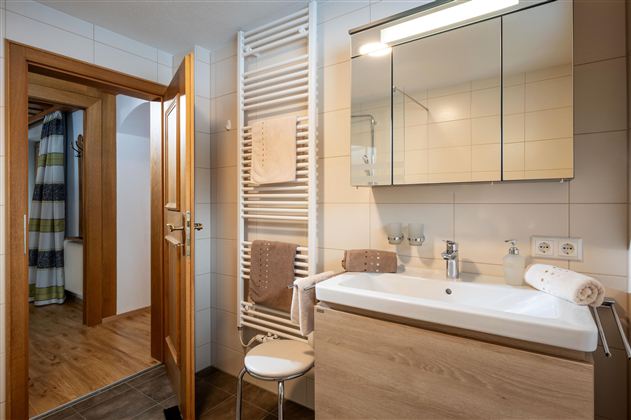 Ein modernes Badezimmer mit einer weißen Waschtischkombination und einem großen Spiegel. Im Hintergrund sind eine Holztür und ein Handtuchhalter zu sehen.