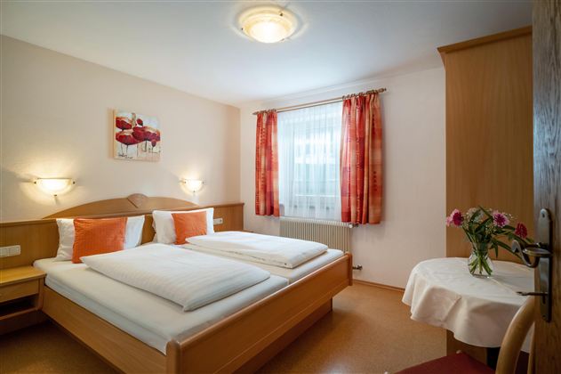 Ein gemütliches Schlafzimmer mit zwei Betten, hellen Wänden und warmen Farben. Neben einem Tisch mit einer Blumenvase gibt es große Fenster mit Vorhängen.