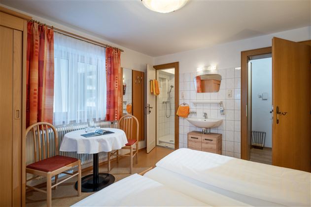 Ein gemütliches Gästezimmer mit zwei Einzelbetten, einem Tisch und Stühlen. Das Zimmer hat große Fenster mit Vorhängen und ein eigenes Bad.