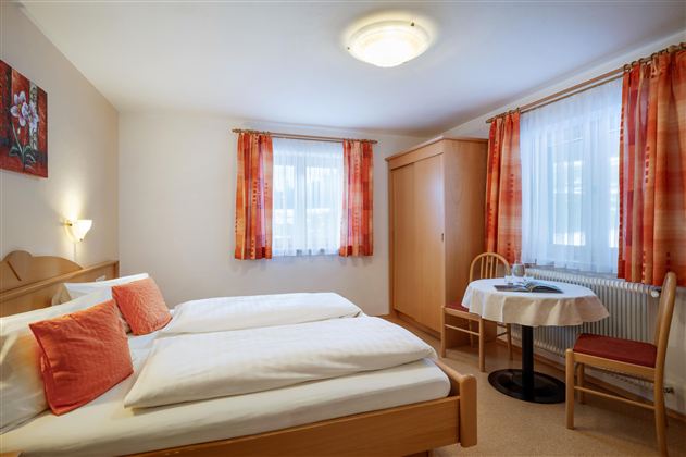 Ein gemütliches Zimmer mit zwei Einzelbetten und einem kleinen Tisch. Die Fenster sind hell und mit Vorhängen dekoriert.