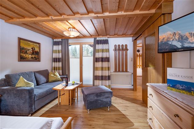 Ein gemütlicher Wohnraum mit rustikalem Holzdekor, einem Sofa und einem TV. Große Fenster sorgen für viel Tageslicht und bieten einen Blick nach draußen.