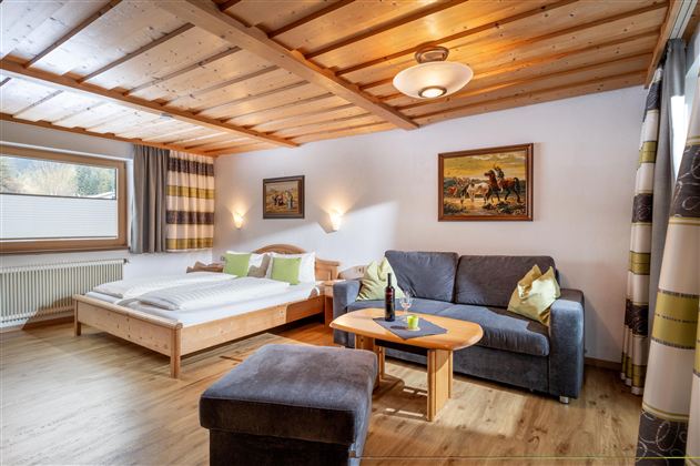 Ein gemütliches Zimmer mit einem Doppelbett und einem Sofa. Die Holzdecke und die warmen Farben schaffen eine einladende Atmosphäre.