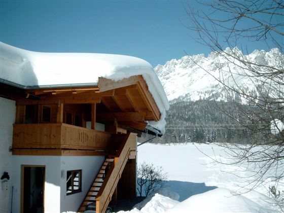 Ein schönes Holzhaus im Schnee mit einem geneigten Dach. Im Hintergrund sind beeindruckende Berge und ein blauer Himmel zu sehen.