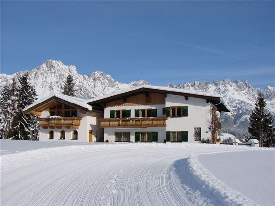 Ein charmantes Haus in den Alpen, umgeben von schneebedeckten Bergen. Der klare Himmel verleiht der winterlichen Landschaft eine friedliche Atmosphäre.