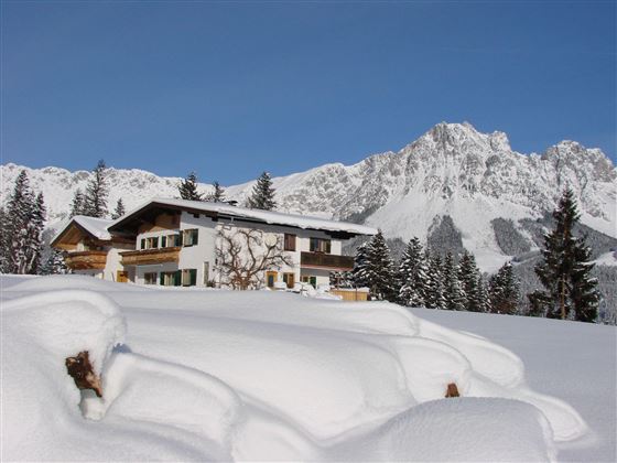 Ein schönes Haus in einer schneebedeckten Landschaft mit Bergen im Hintergrund. Der Himmel ist klar und blau.