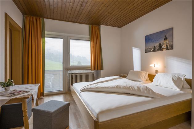 A modern bedroom with a double bed and a cozy wooden ceiling. Large windows let in plenty of light and offer a view of the greenery.