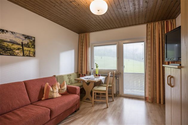 A cozy living space with a red sofa and a dining table. The large windows offer a view of the green landscape.