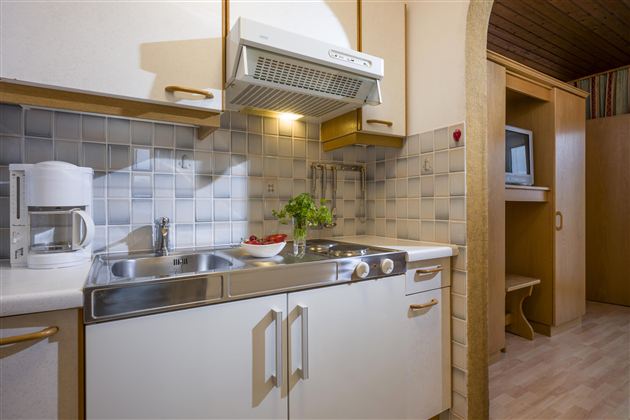 A modern kitchen corner with a sink, stove, and a microwave. The walls are tiled with light tiles and the lighting creates a friendly atmosphere.