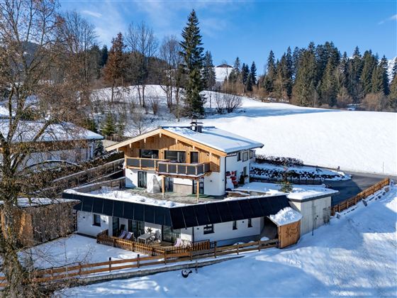 Een met sneeuw bedekt huis in een rustige omgeving. Op de achtergrond zijn hoge bomen en zachte heuvels te zien.