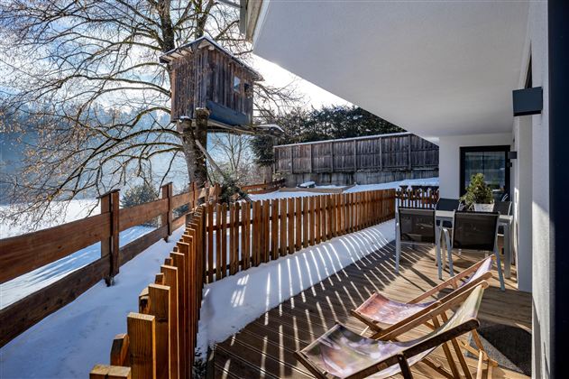 Een gezellige terras met ligstoelen en een houten afscheiding. Op de achtergrond staat een boomhut en de grond is bedekt met sneeuw.