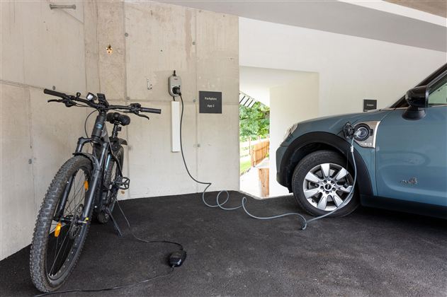 A modern garage environment with an e-bike and an electric car, both of which are charging. On the wall, there is a charging station for electric vehicles.