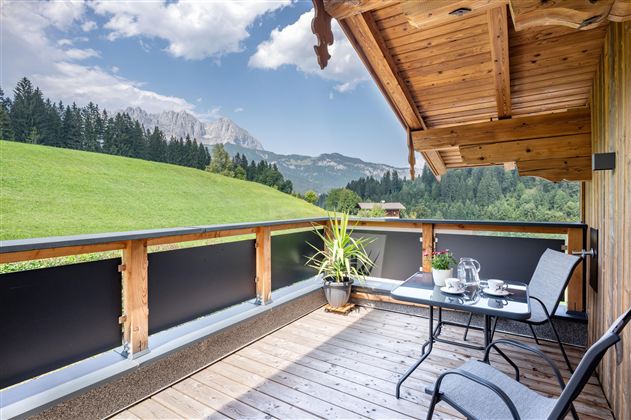 A cozy balcony with a table and chairs. In the background, green hills and mountains can be seen under a blue sky.
