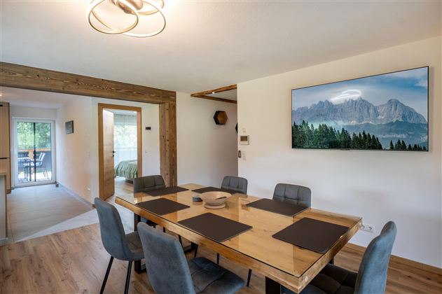 A modern dining room with a large wooden table and six chairs. A picture of mountain landscapes hangs on the wall, while large windows offer a view outside.