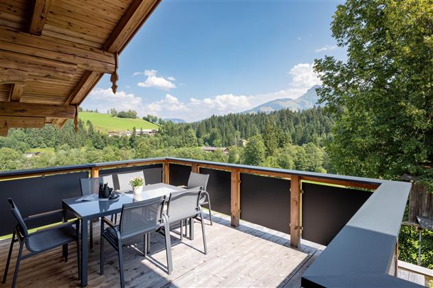A cozy terrace with a table and chairs, surrounded by green forests and mountains. The sky is blue with white clouds.