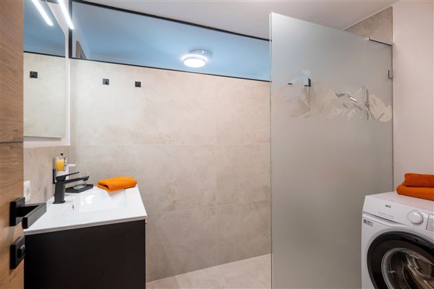 A modern laundry room with a shower and a washing machine. The walls are brightly tiled, and there are orange towels on the countertop.