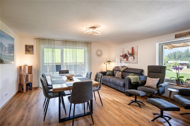 A modern living room with a large dining table and comfortable seating.  
Large windows let in plenty of light and offer a view of the outdoors.