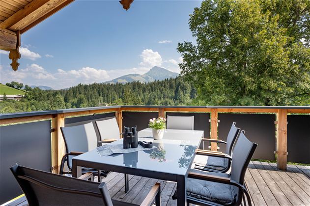 A cozy balcony with a table and chairs, surrounded by green mountains and trees. The sky is blue with some clouds.