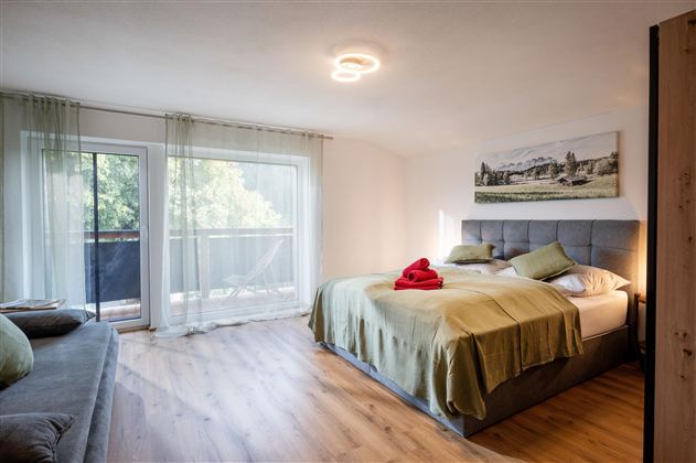 A modern bedroom with a double bed and a cozy sofa. Large windows provide plenty of daylight and a view of the greenery.