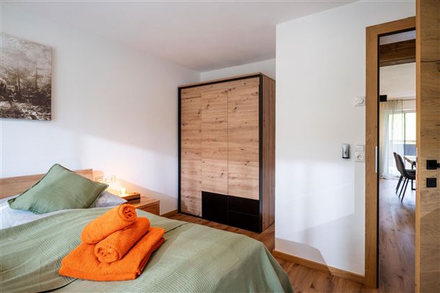 A modern bedroom with a cozy bed and fresh towels. The space is bright and welcoming, featuring a wooden closet and a view of the adjoining room.
