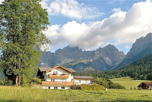 A picturesque house in the midst of nature, surrounded by green meadows and majestic mountains. The sky is partly cloudy, creating a tranquil atmosphere.