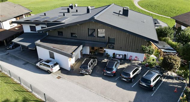 An elegant wooden house with a large roof and colorful flowers in front of the facade. In the foreground, there are parking spaces and a green landscape in the background.