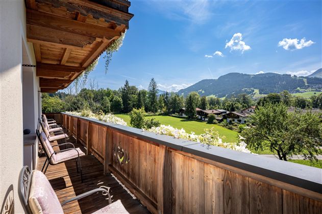 A balcony with a wooden railing and cozy chairs. In the background, there is a green meadow and mountains under a blue sky.