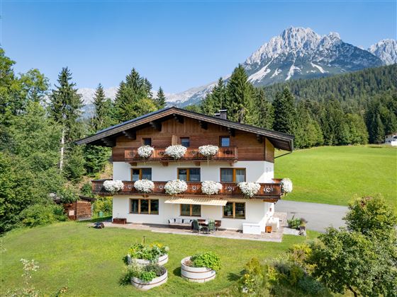 A charming alpine house with flower boxes and a large garden. In the background, majestic mountains and a clear sky can be seen.