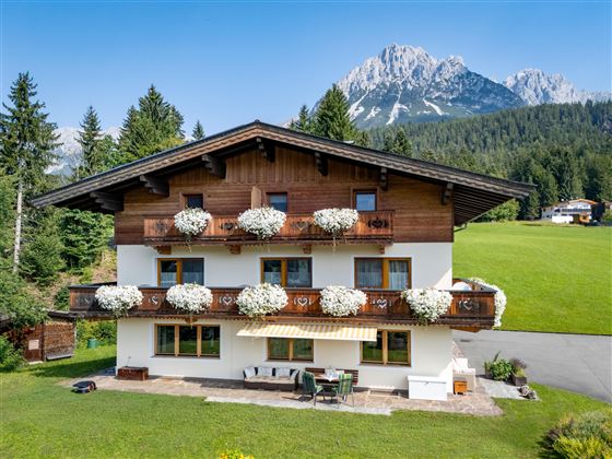 A charming house with a wooden veranda area and blooming flower boxes. In the background, majestic mountains and a green meadow can be seen.