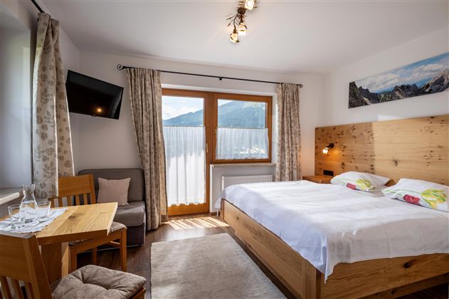 A modern bedroom with a large bed and wooden furniture. There is a table with chairs and large windows that offer a view of the mountains.
