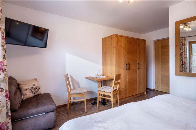 A cozy room with a bed, a table, and two chairs. On the wall hangs a television, and there is a wooden wardrobe.