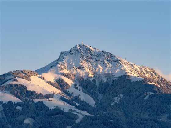 Een majestueuze berg met een met sneeuw bedekte top en zachte hellingen. De lucht is helder en straalt in zachte kleurtinten.
