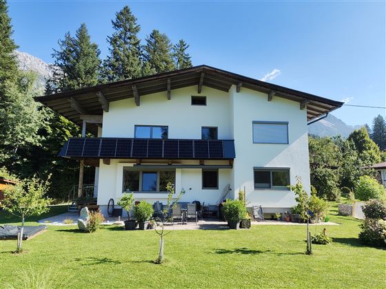 Ein modernes Haus mit Solarpanelen und einem gepflegten Garten. Umgeben von Bäumen und Bergen, bietet es eine schöne Aussicht.
