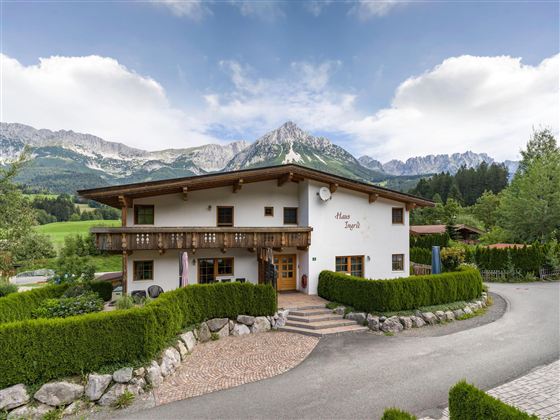 Ein charmantes Haus in den Bergen mit einem gepflegten Garten. Im Hintergrund sind majestätische Berge und ein klarer Himmel zu sehen.