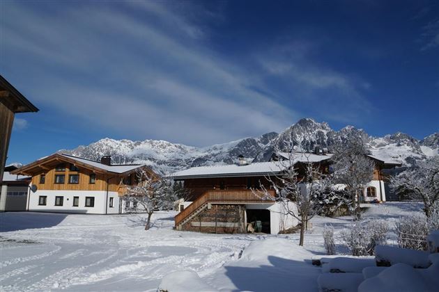 Eine winterliche Landschaft mit schneebedeckten Häusern und Bergen im Hintergrund. Der Himmel ist blau mit einigen Wolken.