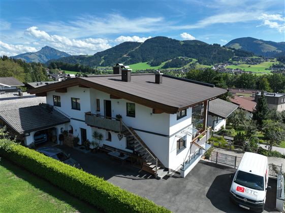 Ein modernes Haus umgeben von grünen Hügeln und Bergen. Der Himmel ist blau mit ein paar Wolken.<br/>Das Haus der Wohnung und im Hintergrund der Blick nach Ellmau.