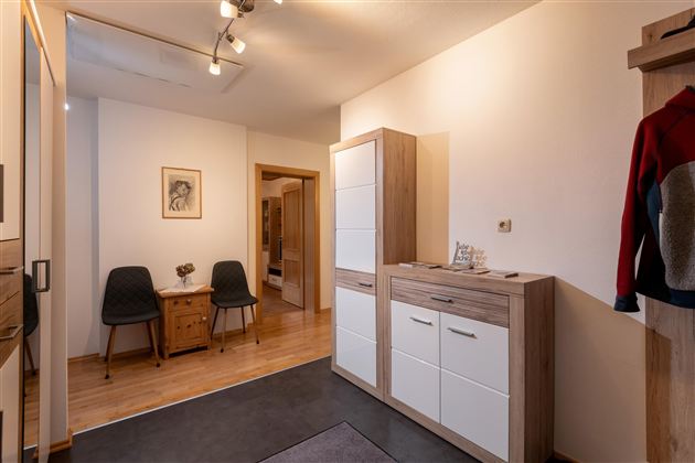 Een moderne hal met een houten vloer en minimalistische meubels. Te zien zijn een commode, stoelen en een mooi wandkunstwerk.