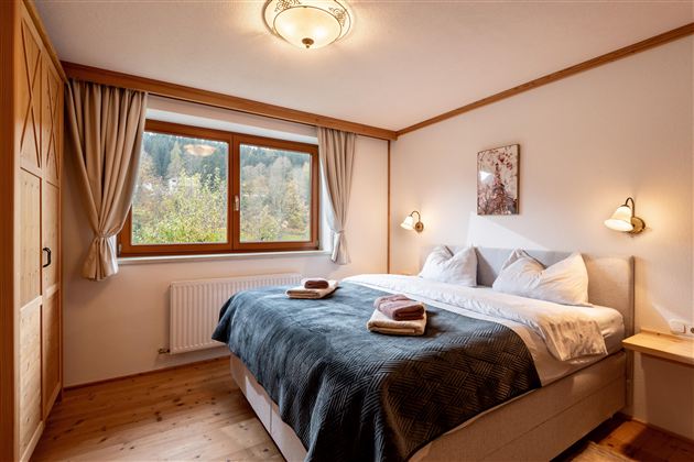 Een gezellige slaapkamer met een groot bed en een raam dat uitkijkt op de natuur. Het is helder en uitnodigend, met een warme houten plafond en zachte kleuren.