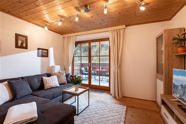 Een gezellige woonkamer met een grijze bank en een tafel. Grote ramen met gordijnen bieden uitzicht op een balkon.