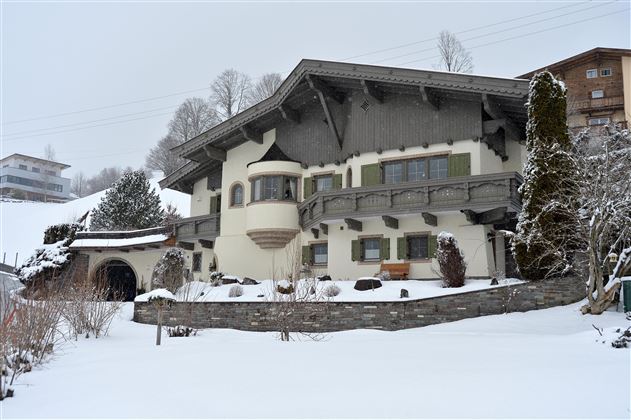 A charming alpine house in the snow, surrounded by snow-covered mountains. The architecture is traditional with a charming bay window and wooden embellishments.