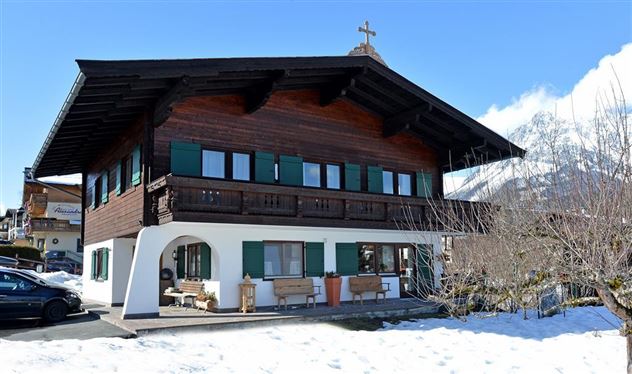 Een traditioneel houten huis met groene shutters en een kruis op het dak. Het staat in een met sneeuw bedekt landschap met bergen op de achtergrond.