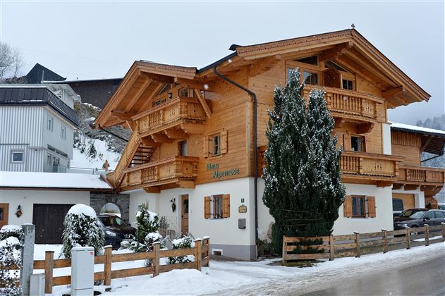 Een mooi houten huis in alpine stijl met meerdere balkons. Er ligt sneeuw rondom en de sfeer is gezellig.