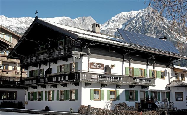 Ein traditionelles alpenländisches Haus mit einem schönen Balkon und grünen Fensterläden. Im Hintergrund sind schneebedeckte Berge zu sehen.