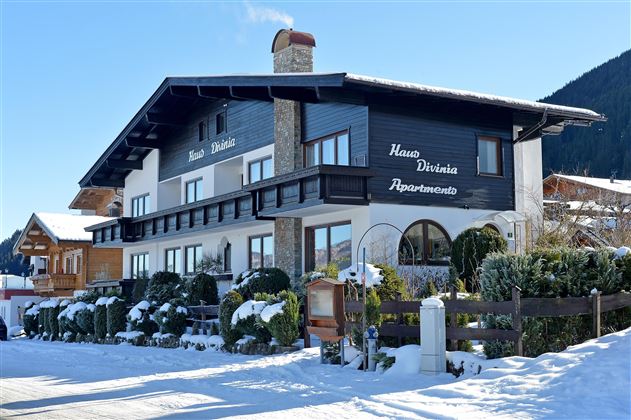A charming building in alpine style, surrounded by snow and well-kept plants. It offers cozy apartments and an inviting entrance area.