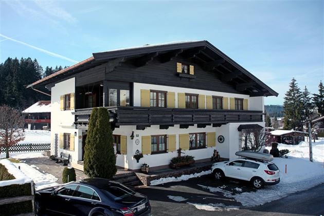 Ein traditionelles alpenländisches Haus mit schwarzem Holz und gelben Fensterläden. Es liegt in einer verschneiten Umgebung mit Bäumen im Hintergrund.