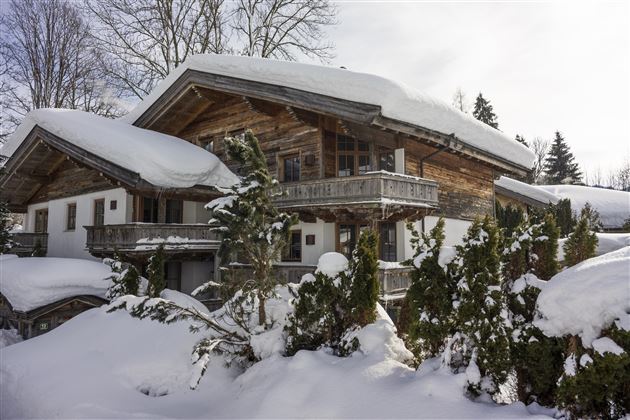 A rustic wooden house in the snow, surrounded by tall coniferous trees. The sky is clear and a peaceful winter landscape can be seen.