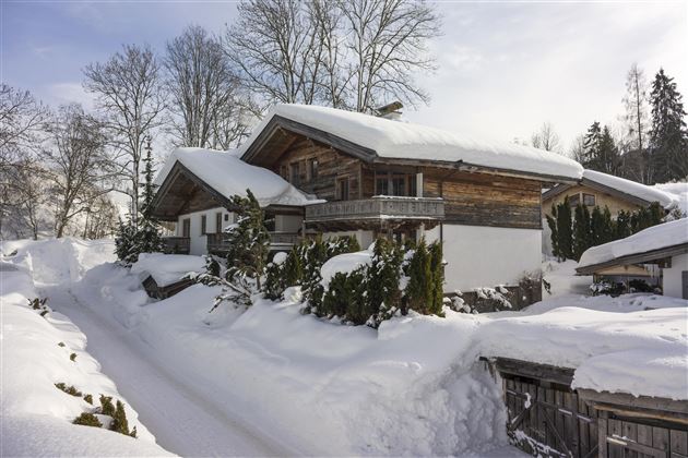 A cozy wooden house in the snow, surrounded by trees. The landscape is wintry and peaceful.