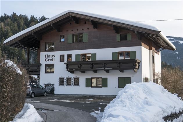Ein gemütliches Haus im alpinen Stil, umgeben von Schnee. Die Fenster sind grün und das Dach ist überhängend.