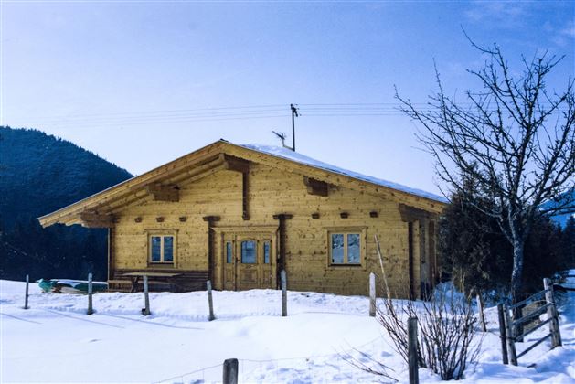 Ein gemütliches Holzhaus in einer verschneiten Landschaft. Im Hintergrund sind Berge und blauer Himmel zu sehen.